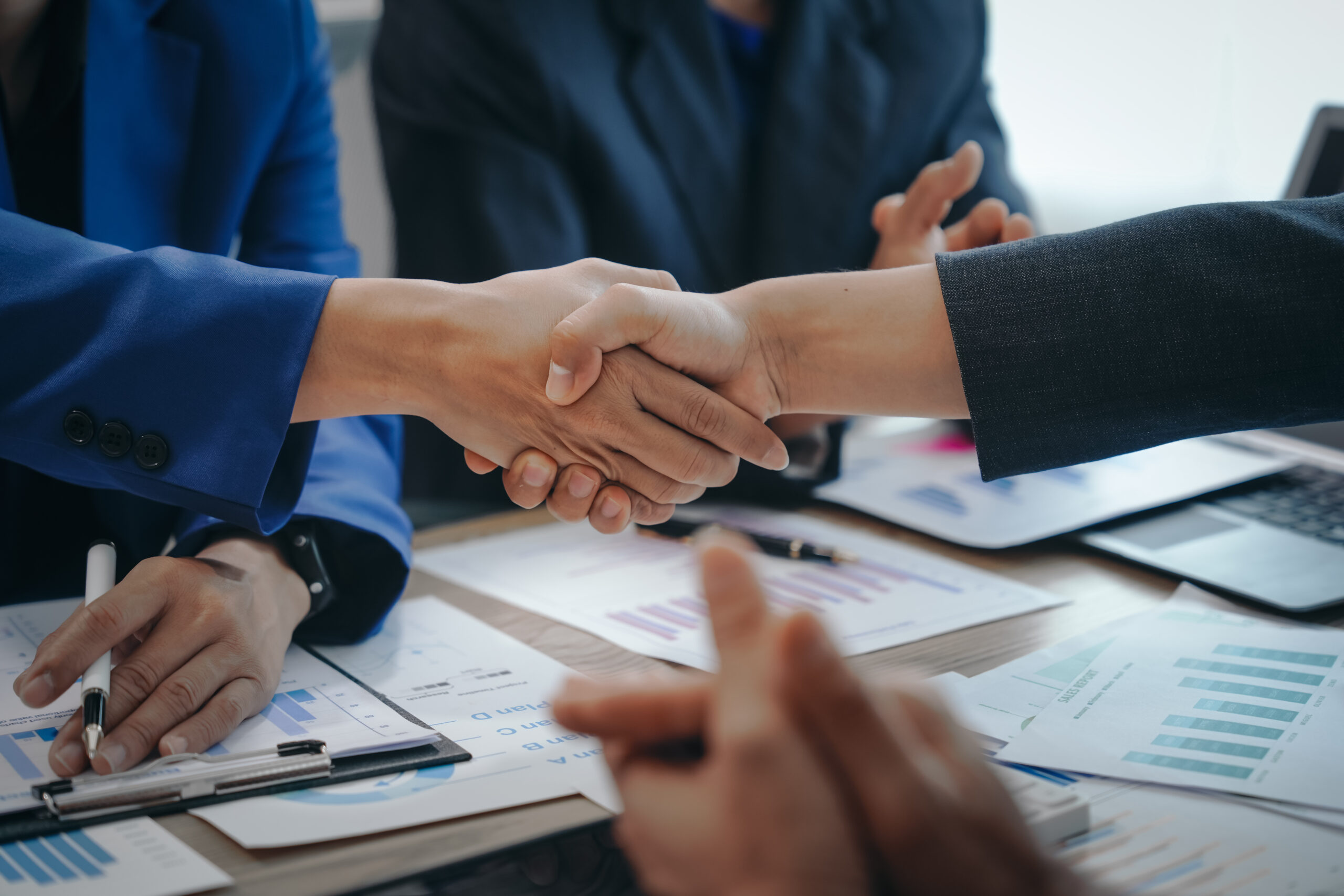 A stock image of a business team meeting, success is celebrated with a handshake. Hands close up, dressed in formal suits, working at desks with financial papers, calculators, and laptops.