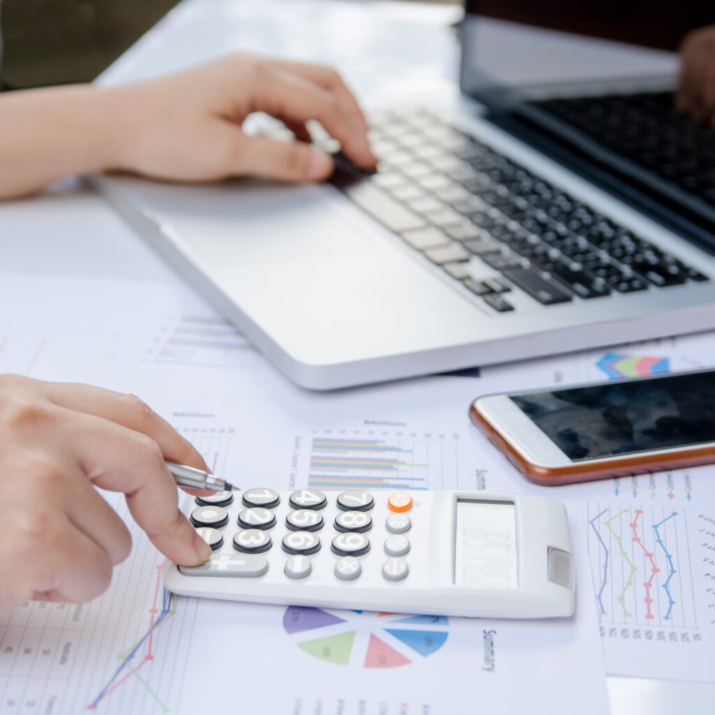 A close up of a woman using a calculator and laptop on paper with graph data whilst doing finance at the office.