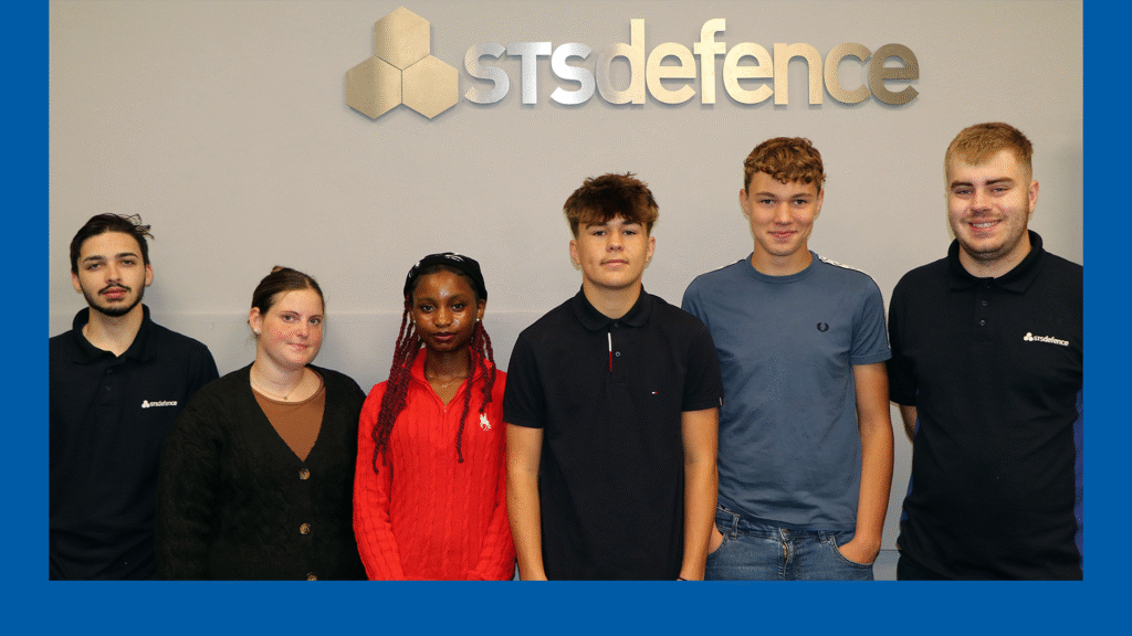 A group photo of STS Defence apprentices in a line against a grey wall, with the STS Defence logo above their heads.
