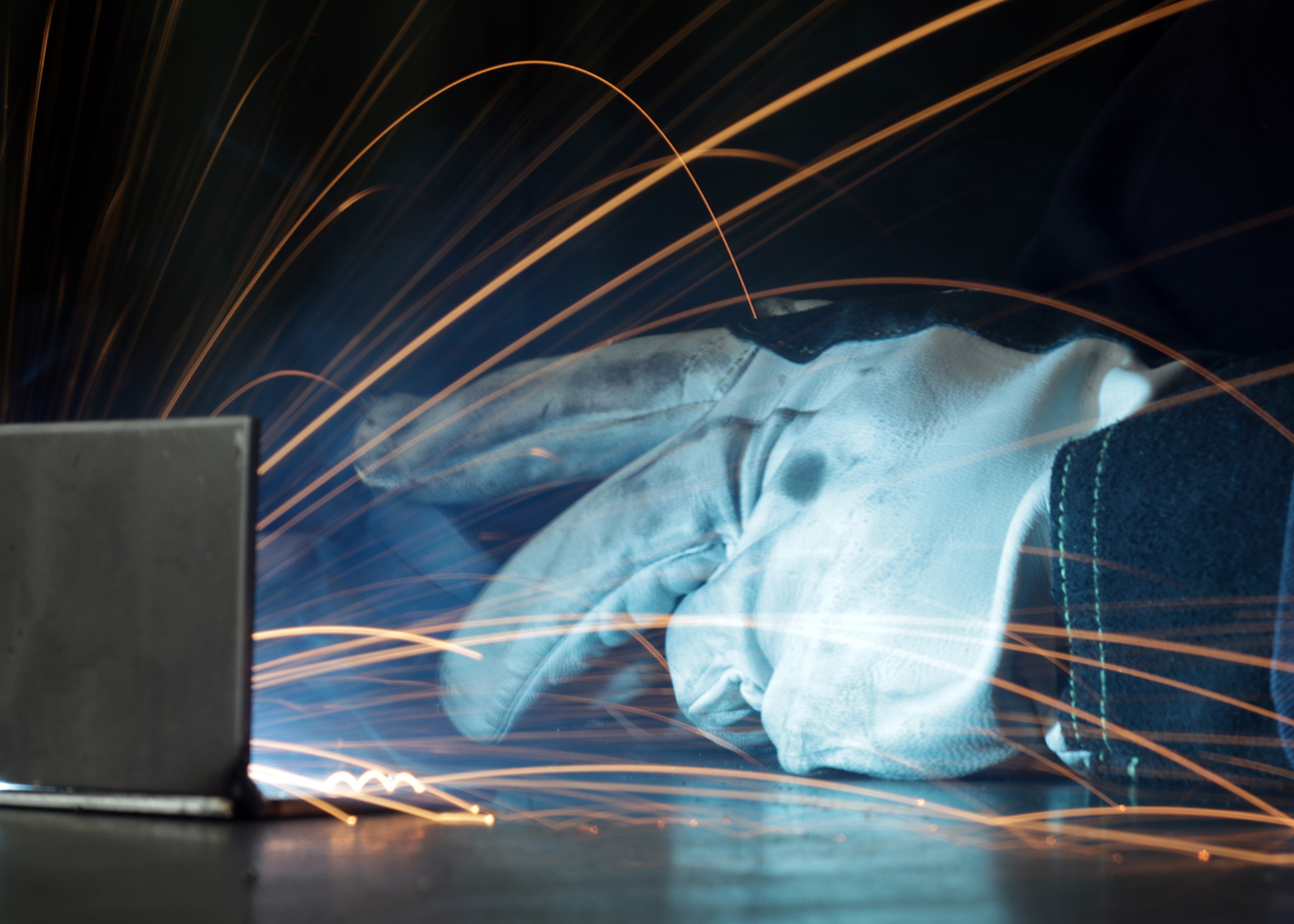 A photo of a welder's gloved hand working on a piece of metal with the sparks captured.