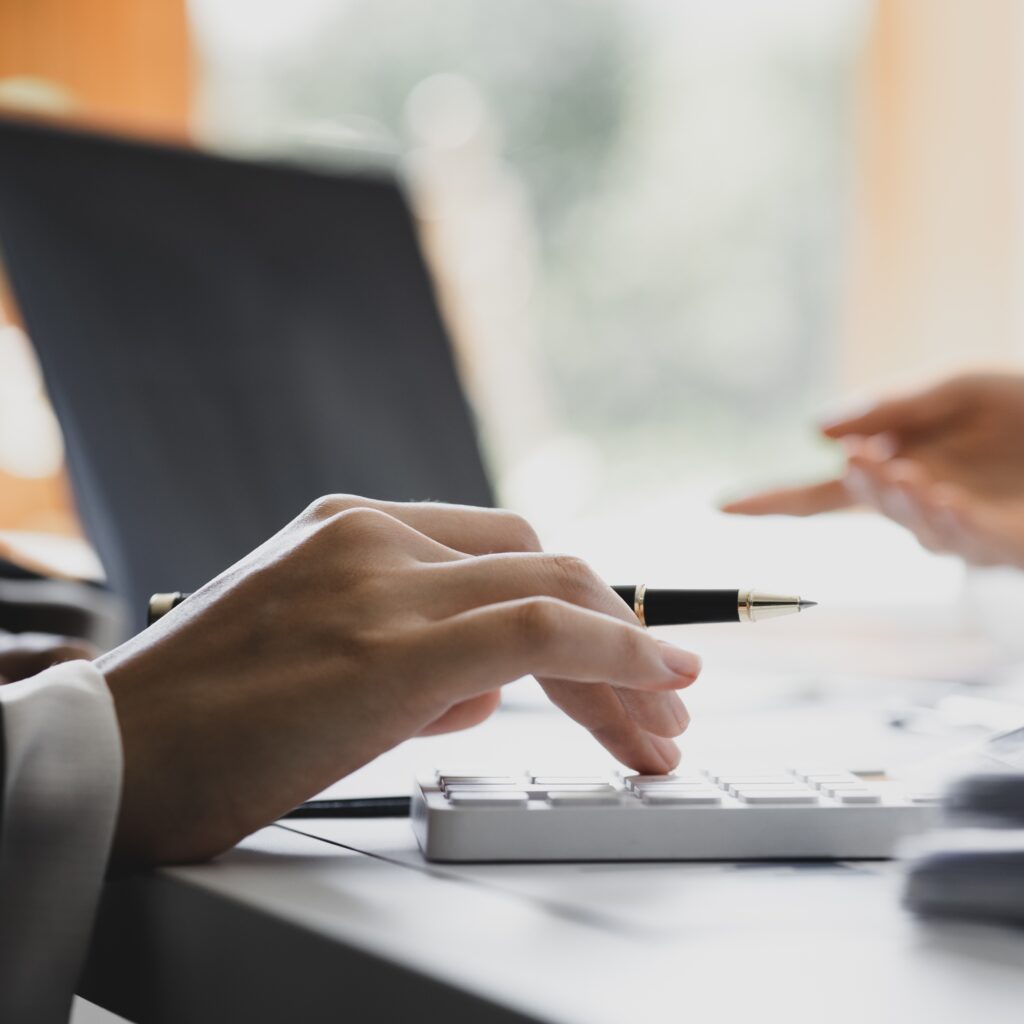 A cropped stock image of two people having a meeting. One is holding a pen and there is a laptop in the background.