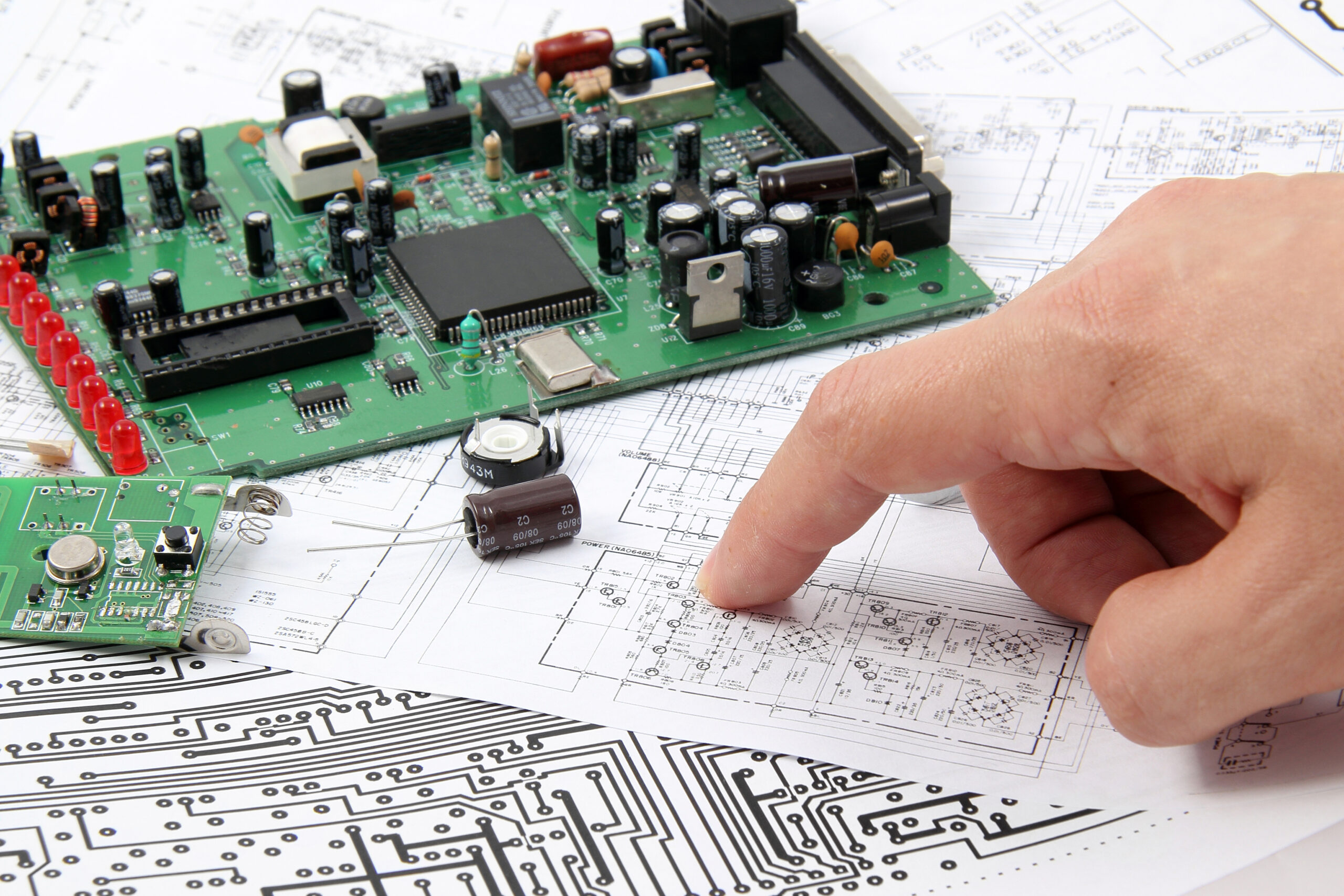 A stock image of a hand pointing at a diagram of a circuit board, with an incomplete circuit board nearby.