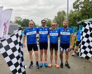 A photo of the 2025 STS Defence Extra Mile Challenge team. There is a group of four men in blue branded cycling shirts at the finish line with black and white checked flags either side of them.