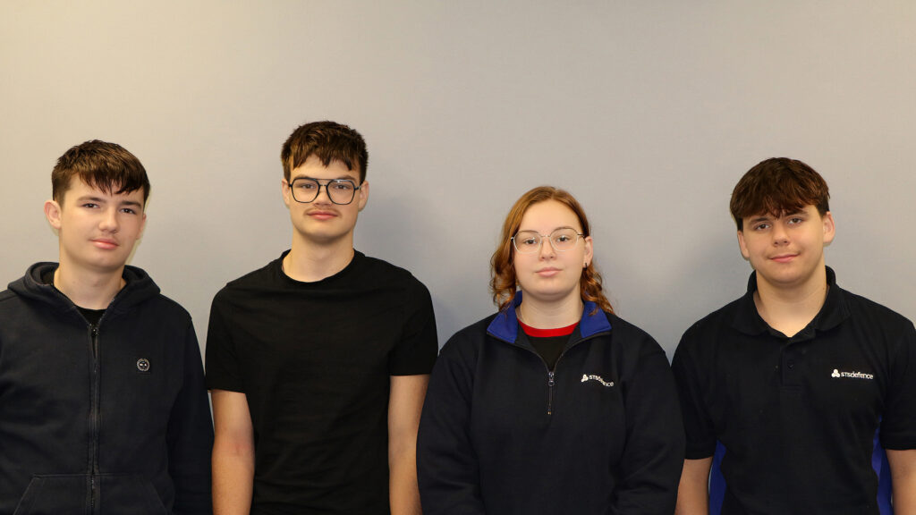 A group of four STS Defence apprentices stood against a grey wall facing the camera